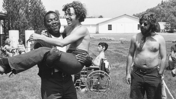 a screenshot from the film Crip Camp shows a white disabled man being carried by black disabled man outdoors in the camp in upstate New York in the 70s. There are other disabled people in the background including a man in a manual wheelchair. The photo is black-and-white.
